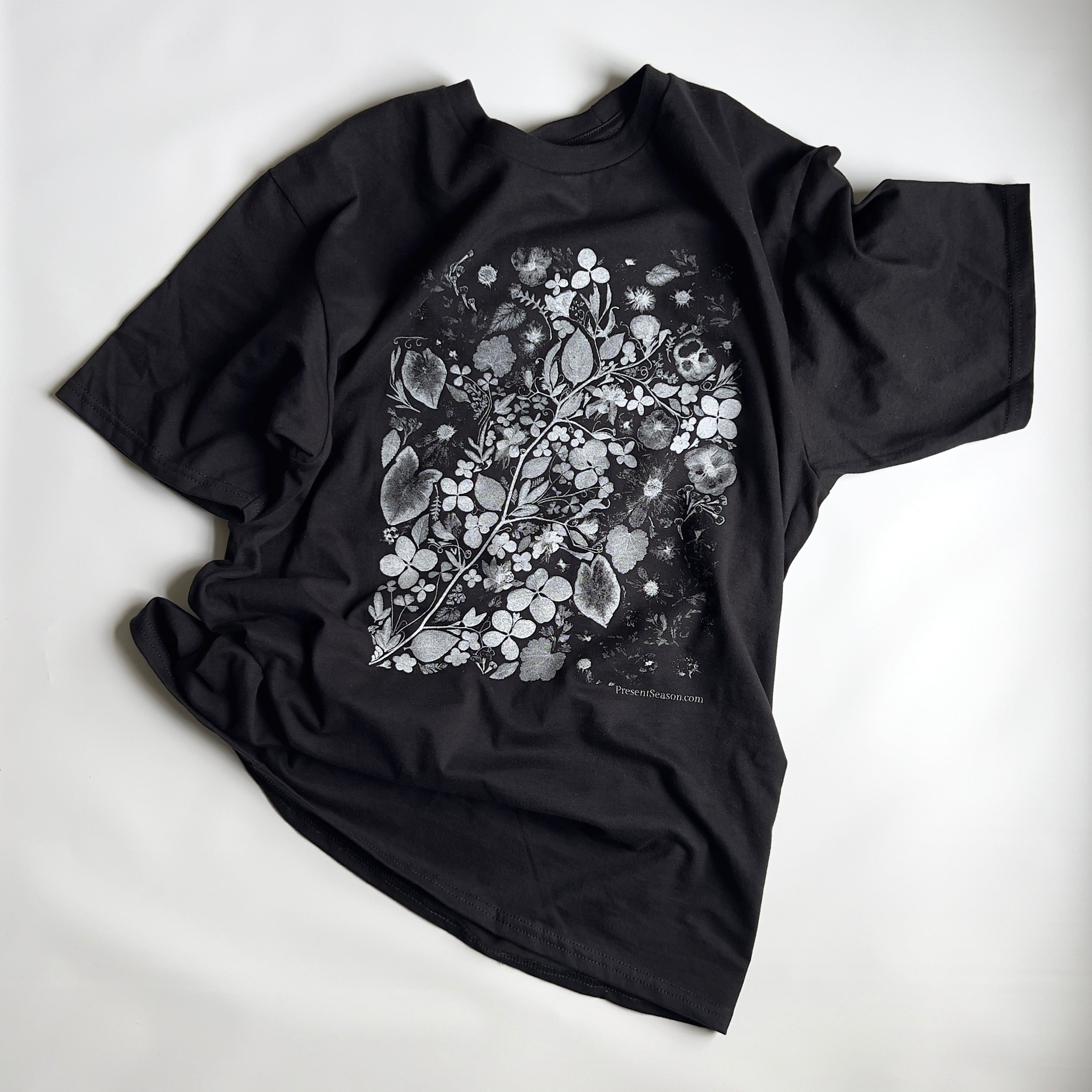 A black short sleeved t-shirt with a white and gray rectangular print of an art scene made from pressed flowers.