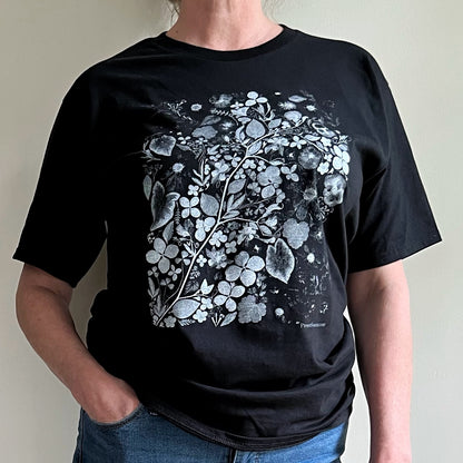 A person wearing a black tshirt with a white and gray flower print on it. The person has their hand in one pocket and is wearing jeans. 