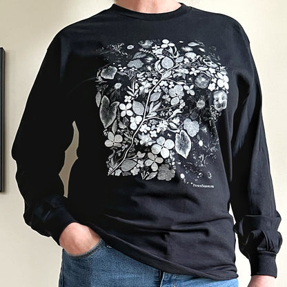 A person is wearing a black long sleeve tshirt and has one hand in their jeans pocket.  The screen print on the t-shirt is white and gray and is flowers and leaves. 