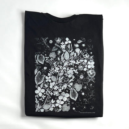 a black are t-shirt on a white background. The shirt is folded into a rectangle with just the sides and collar showing. There is a white and gray art print on the shirt and it has been created with many flowers and houseplant leaves. 