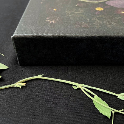 The corner and side of a very thick art piece is covered in black canvas. You can see some fain pressed flowers on the top of the canvas.. It is upon a piece of black fabric. There is a green pressed vine and a few leaves at the bottom of the photo.