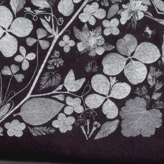 A short video of a close-up of a black fabric t-shirt with a flower print on it. The print is white and gray and consists of many artfully designed flowers and plants. It is on a white background. 